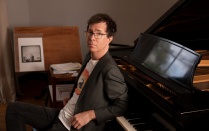 ben folds sitting on a piano bench leaning back on a grand piano with elbows on the keys. 