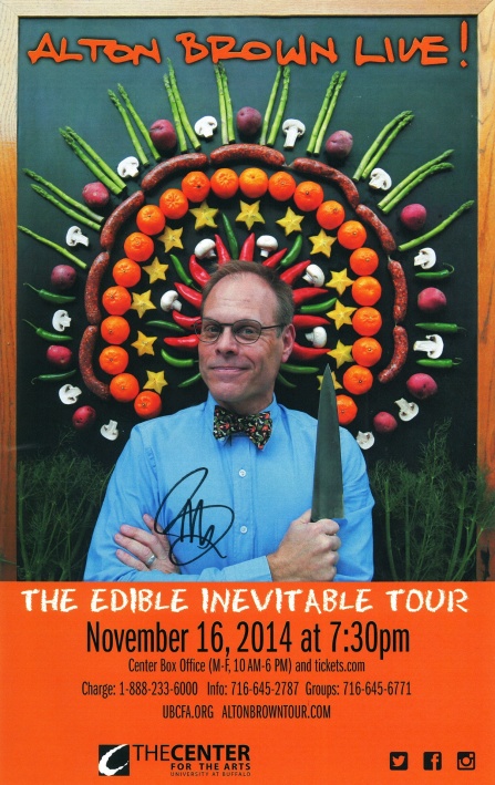 alton brown posing with a chef knife in front of a mosaic of fruits and vegetables.