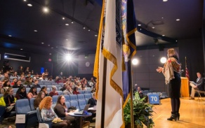 A presenter speaks on stage with a crowd in the background. 