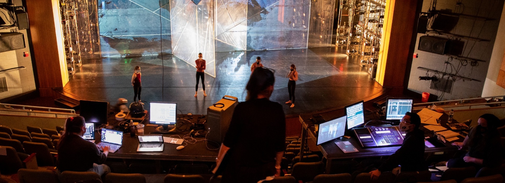 Stage with rehearsal and students working at control and sound board computers from up in the seating. 
