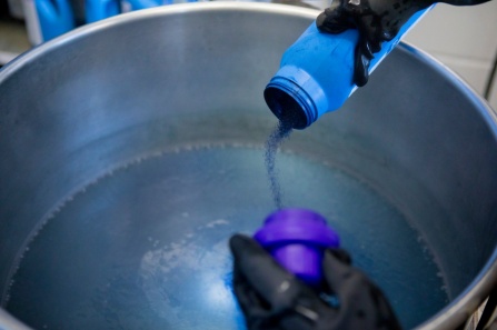 Close up of hands, wearing rubber gloves, pouring liquid into a large pot. 