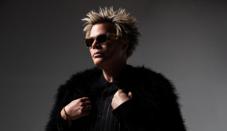 Brian Culbertson wearing glasses and a black fur coat in front of a blank background. 