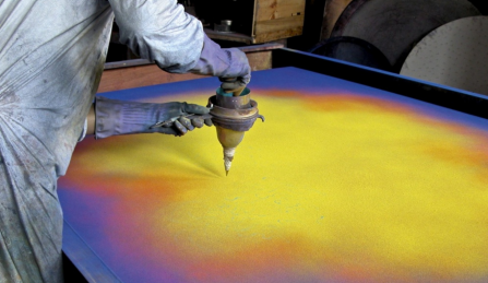 Natvar Bhavsar leans over a large canvas laid flat on a table, sifting dry pigment from a metal funnel onto its surface. The canvas glows with fields of yellow, orange, and violet. The artist wears a pigment-dusted smock and long rubber gloves. 