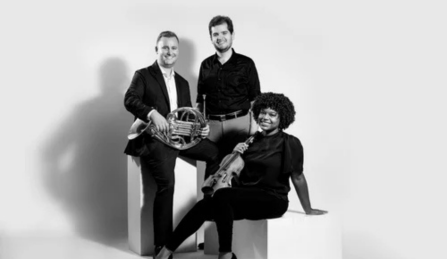 Members of Black Moon Trio posing sitting and leaning on blocks in front of a black background. 