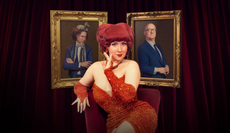 Lucy Darling sitting in a velvet chair in front of a theatre curtain and two hanging portraits. 