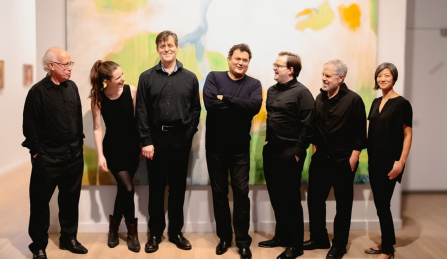 Members of NYNME standing together all in black in front of a large painting. 
