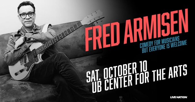 fred armisen sitting on a couch holding an electric guitar. 