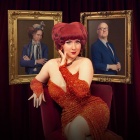 Lucy Darling sitting and posing in front of framed pictures and a velvet curtain. 