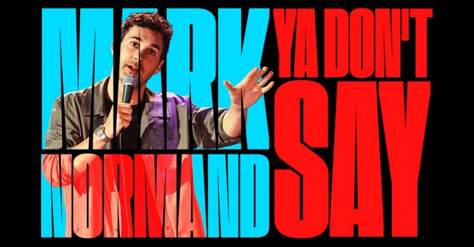 mark normand holding his hand up while talking into a microphone. 